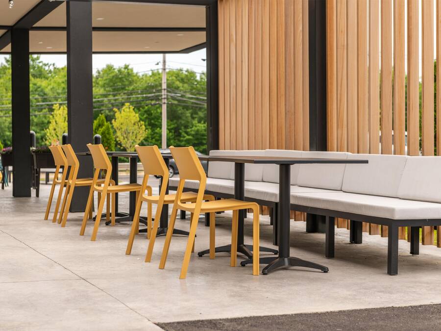 Un área de estar moderna al aire libre con una fila de sillas amarillas y un sofá blanco, suelo de hormigón y un muro de listones de madera al fondo.