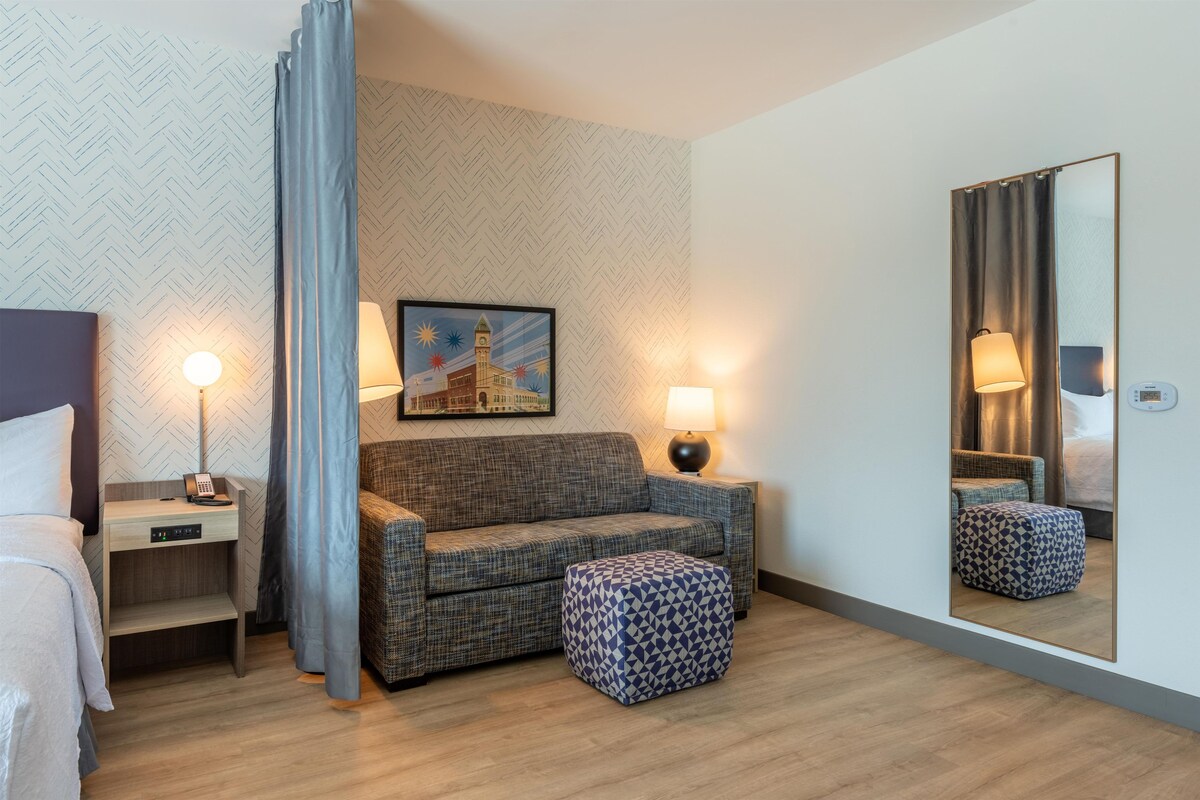A hotel room with a bed, a sofa, a small table, a lamp, a mirror, and a picture on the wall.
