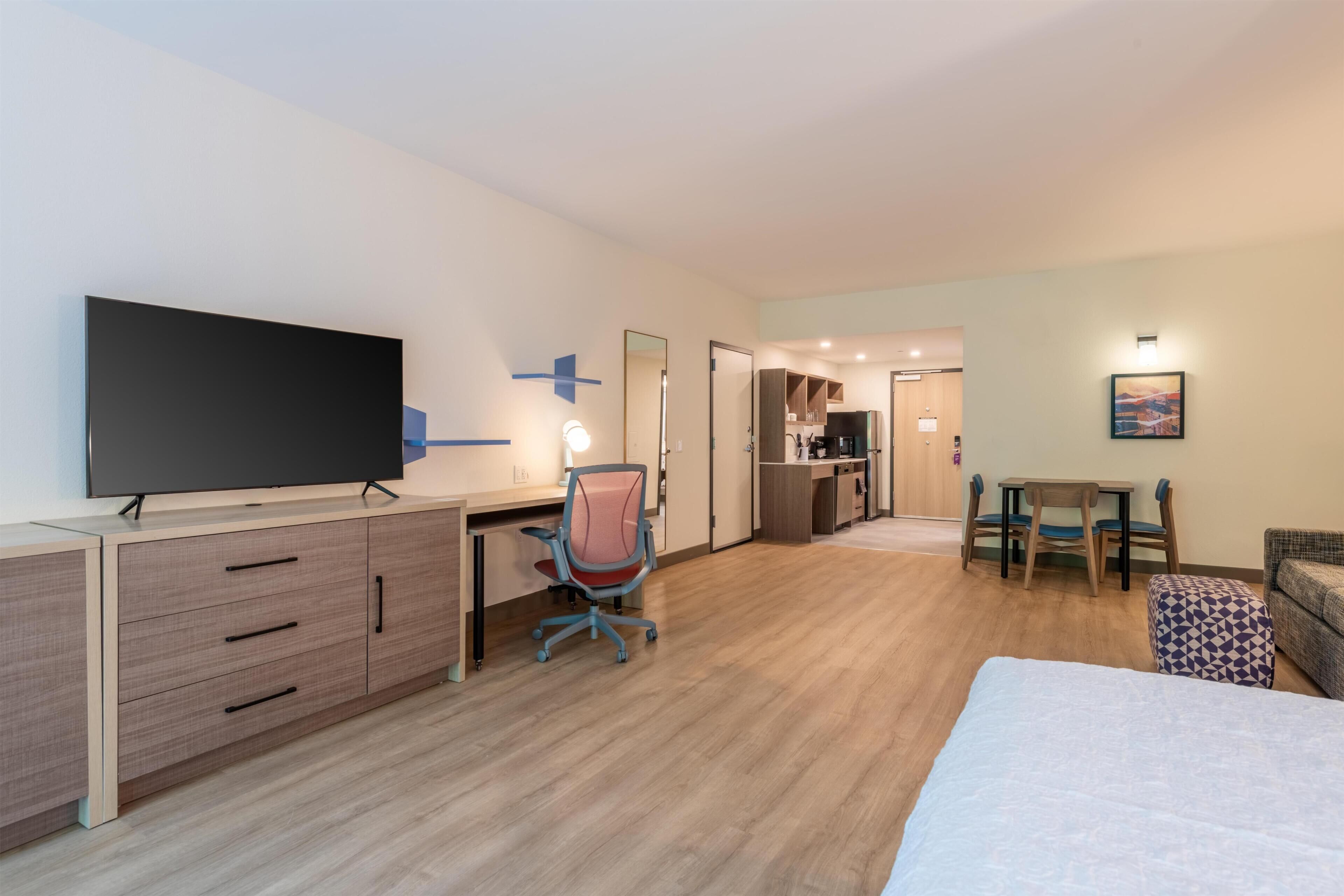 A hotel room with a bed, a desk with a flat-screen TV, a chair, a dining table with chairs, and a kitchenette.
