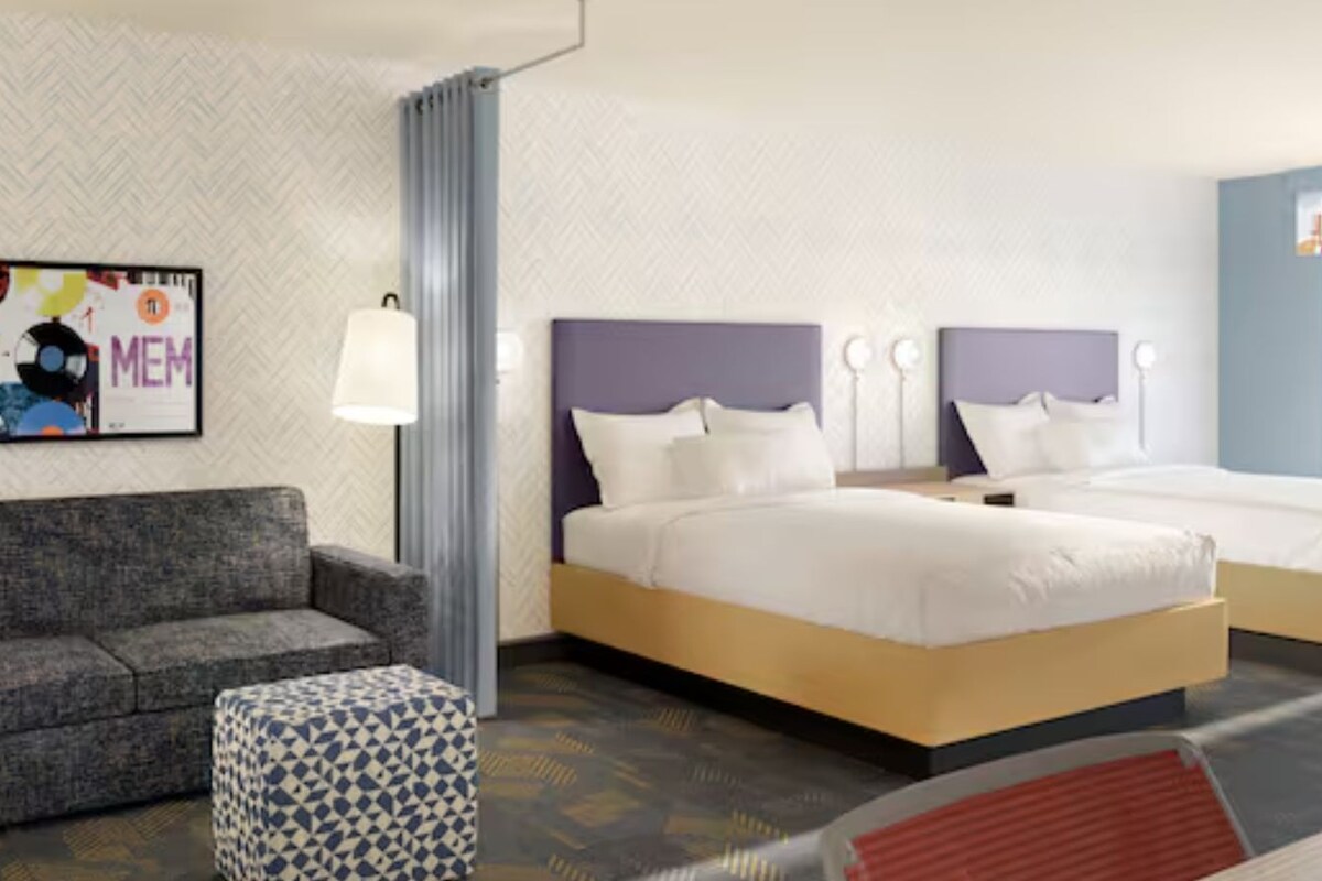 Habitación de hotel con sofá, dos camas, un reposapiés estampado, una pared con un cartel y una ventana con cortinas.