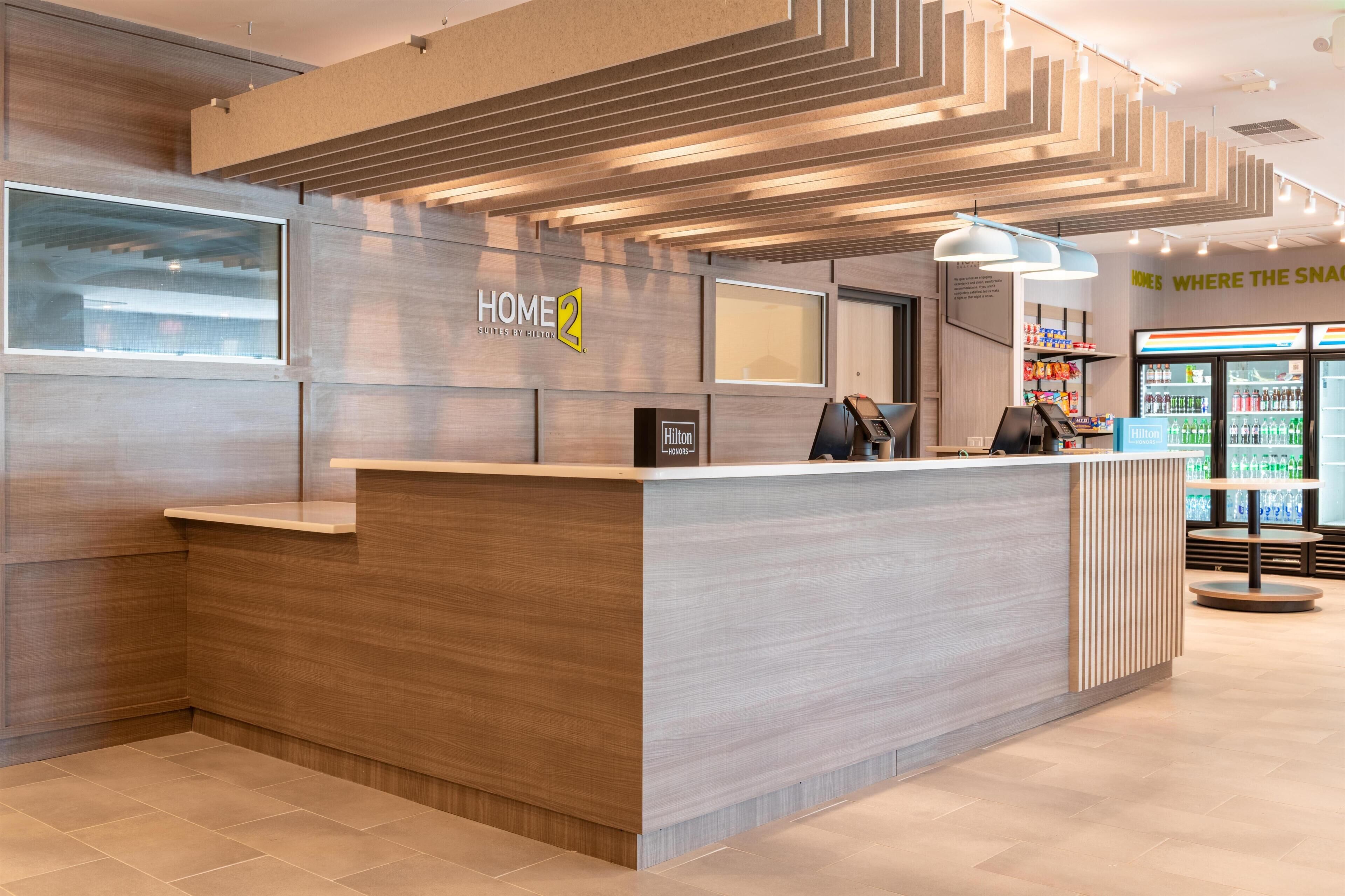 Un comptoir d’accueil moderne, avec une enseigne « HOME 2 », un minibar et un kiosque 7-Eleven.