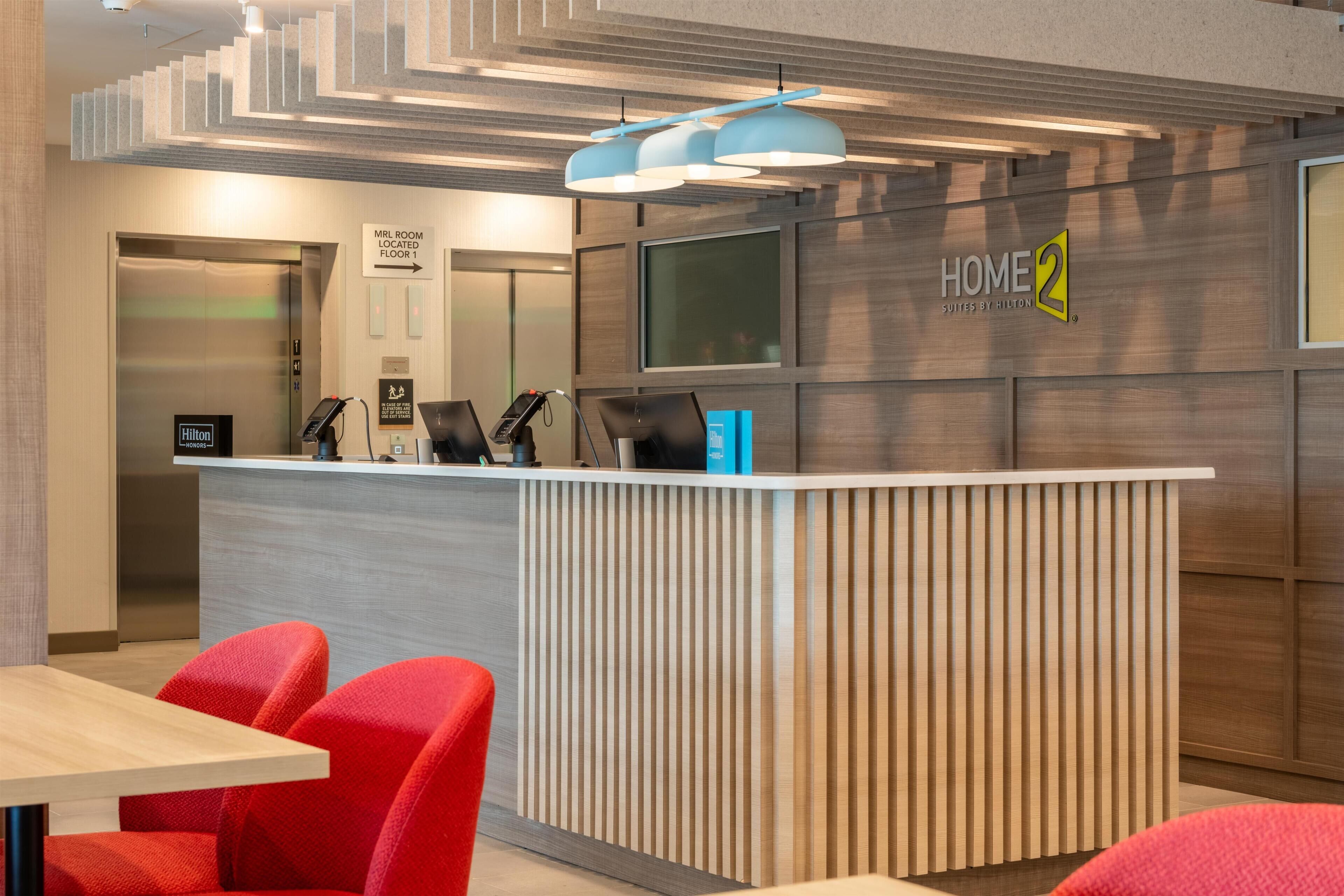 Un área de recepción de hotel moderna, con un mostrador de madera, sillas rojas y un cartel que indica la ubicación de la sala MRL en el primer piso.