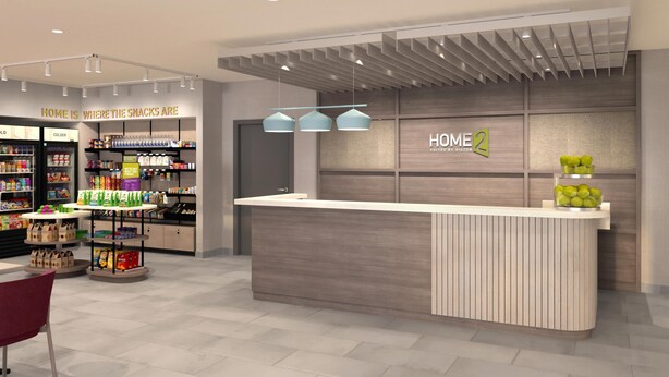 Un área de recepción de hotel moderna con un mostrador de madera, una exhibición de aperitivos y un cartel que dice "HOME 2 SUITES BY HILTON".