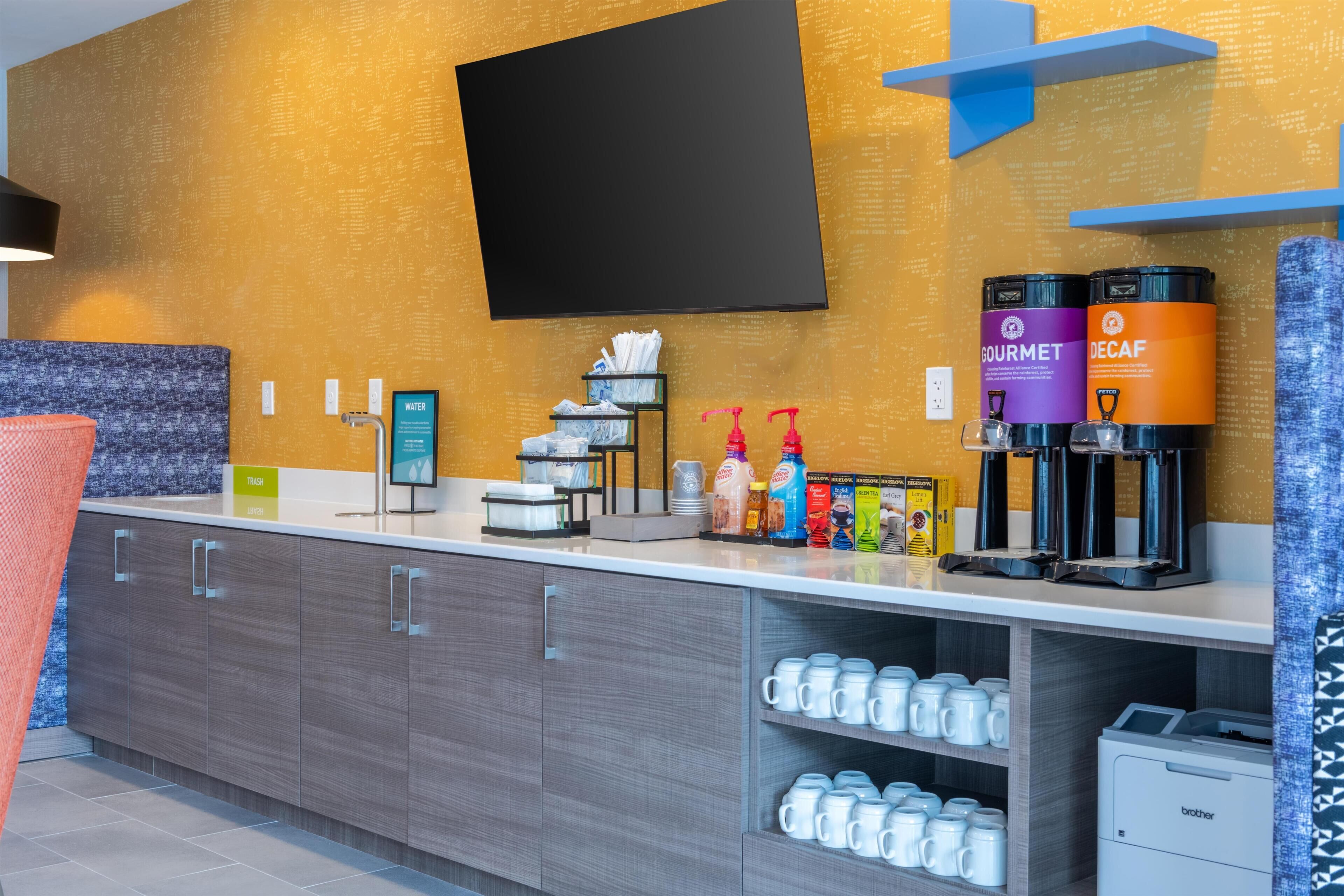 A modern hotel room with a flat-screen TV mounted on the wall, a coffee station with branded dispensers, and a counter with a trash bin and a water dispenser.