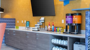 A modern hotel room with a flat-screen TV mounted on the wall, a coffee station with branded dispensers, and a counter with a trash bin and a water dispenser.