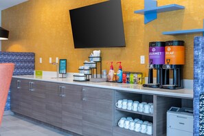 A modern hotel room with a flat-screen TV mounted on the wall, a coffee station with branded dispensers, and a counter with a trash bin and a water dispenser. - Home2 Suites By Hilton North Attleboro (North Attleboro)