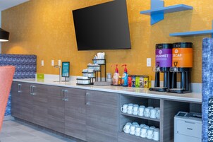 Una habitación de hotel moderna con una televisión de pantalla plana montada en la pared, una zona de café con dispensadores de la marca y una encimera con un cubo de basura y un dispensador de agua.
