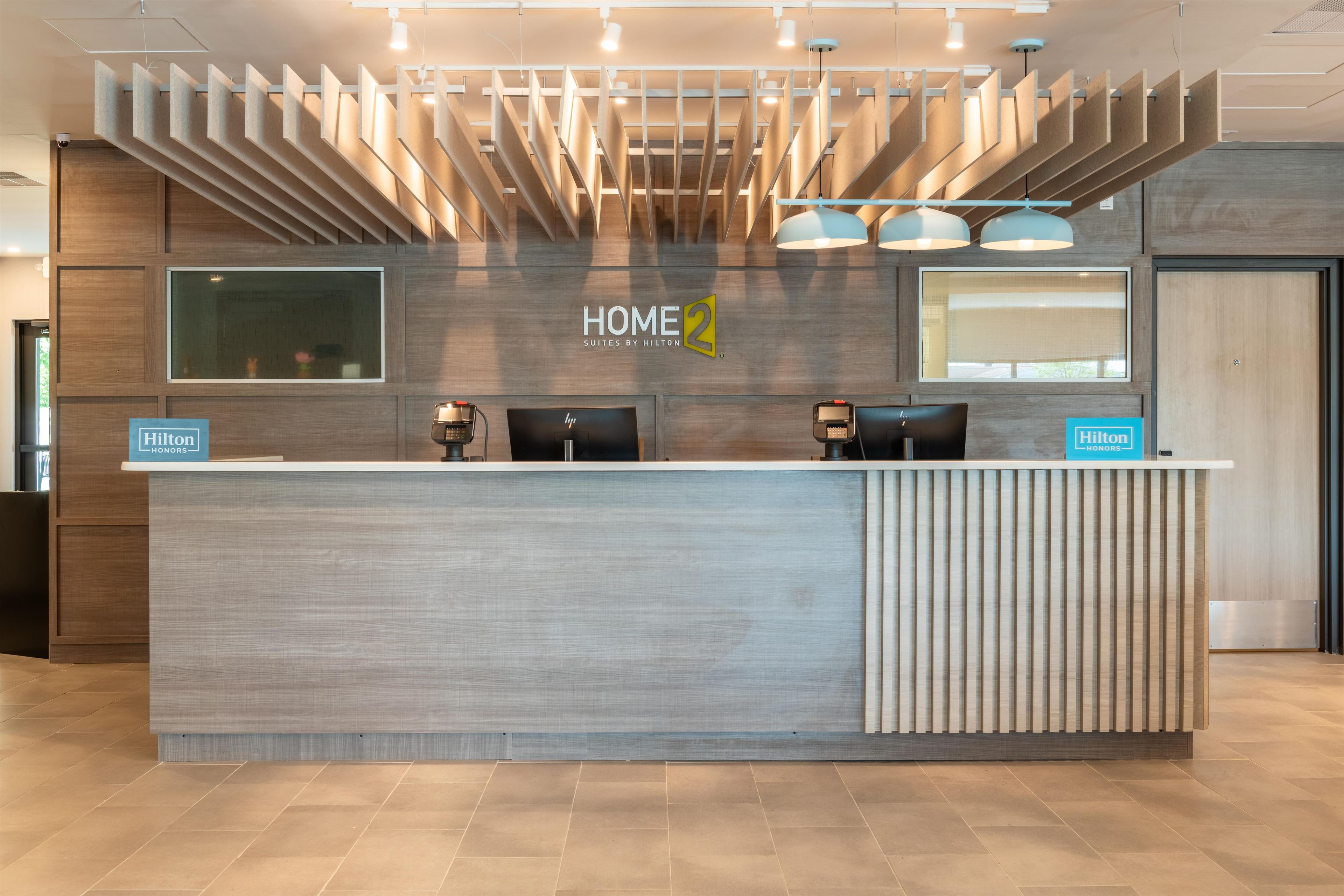 A modern hotel reception desk with a 'HOME 2' sign, two black chairs, and a 'Hilton Honors' sign.