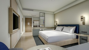 Ein modernes Hotelzimmer mit einem großen Bett, einem Fernseher, einem Badezimmer mit Waschbecken und Spiegel sowie einem kleinen Tisch mit einer Zeitschrift.