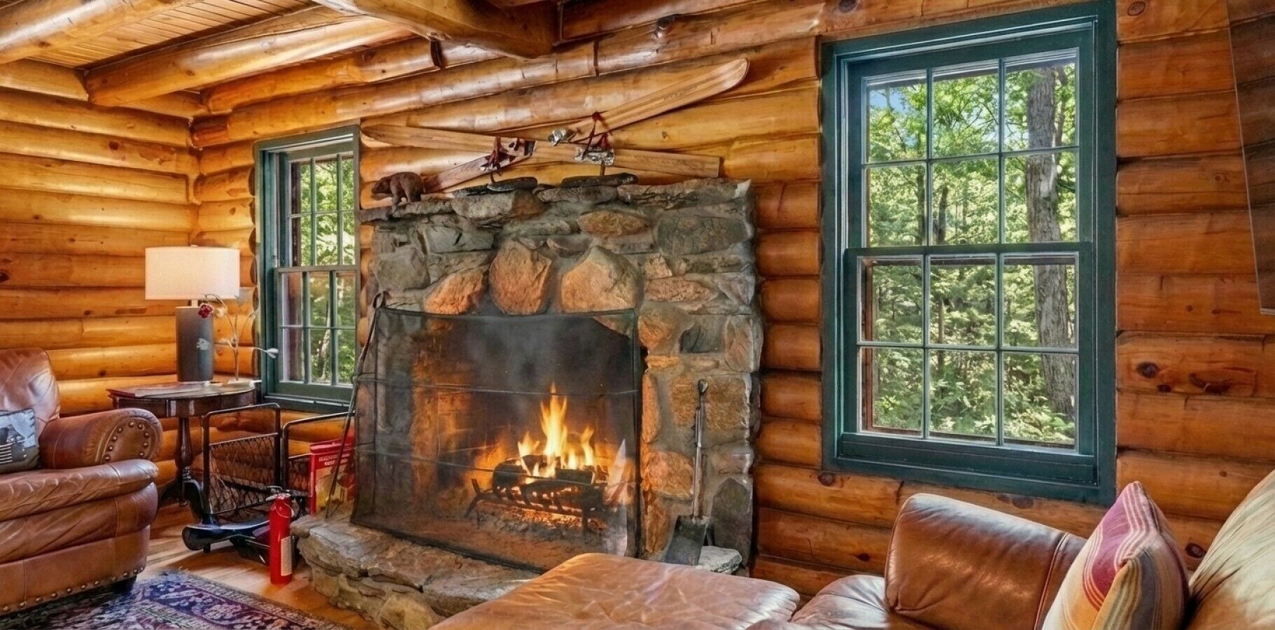 A log cabin interior with a stone fireplace, leather furniture, and large windows.