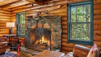 A log cabin interior with a stone fireplace, leather furniture, and large windows.