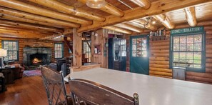 A log cabin interior with a stone fireplace, wooden dining table, and a view of the outside.