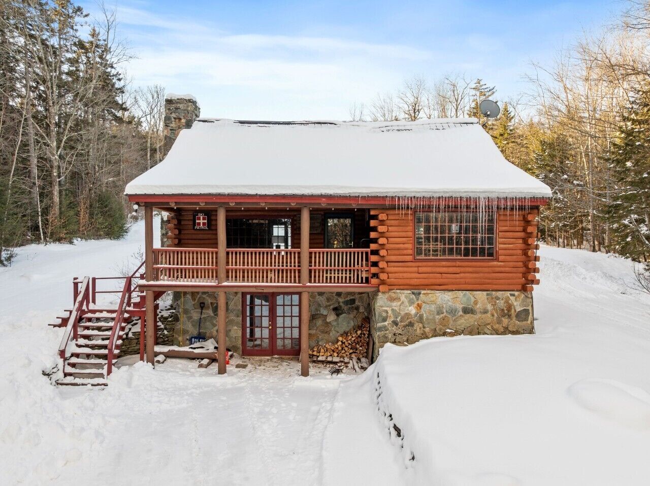 A log cabin with a porch, a stone foundation, and a snow-covered roof.