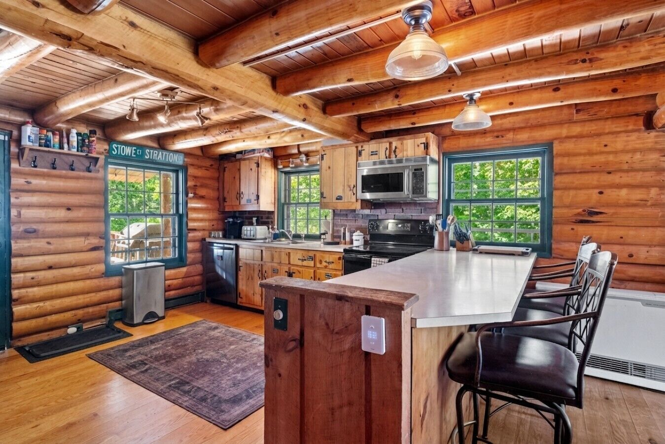 A log cabin kitchen with wooden walls, a central island, bar stools, a microwave, stove, and a large window.