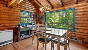 Interior de uma cabana de troncos com uma mesa de jantar, cadeiras, um lustre e grandes janelas com vista para o verde.