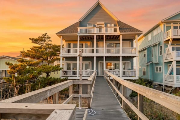 A multi-story building with a balcony on the second floor, a wooden walkway leading to the entrance, and a clear sky in the background. - Smile Maker by Carolina Getaway - Oceanfront w/ Elevator, Sleeps 25 (Carolina Beach)