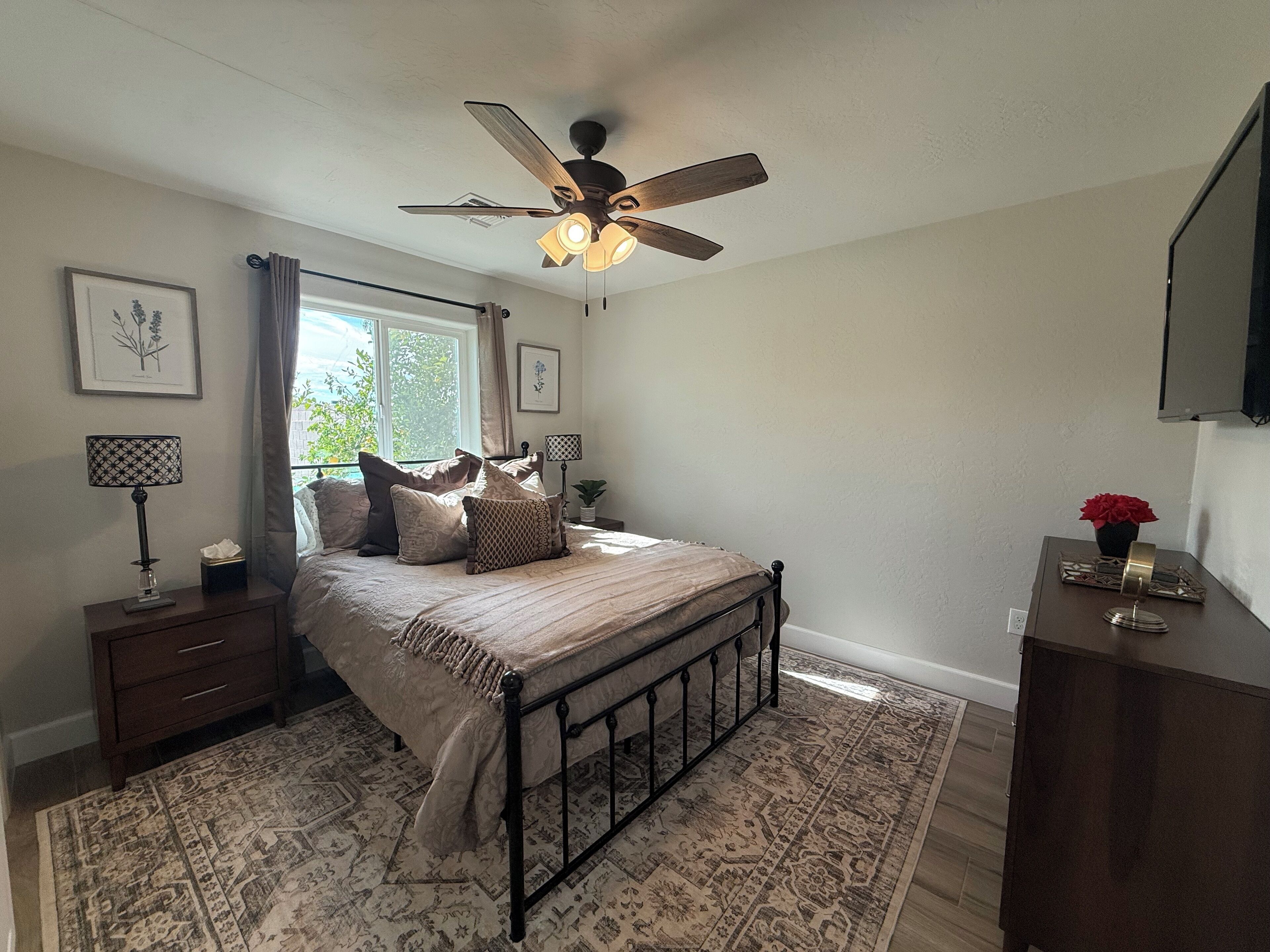 A bedroom with a bed, a ceiling fan, a nightstand, a lamp, a window with curtains, a framed picture, and a potted plant.