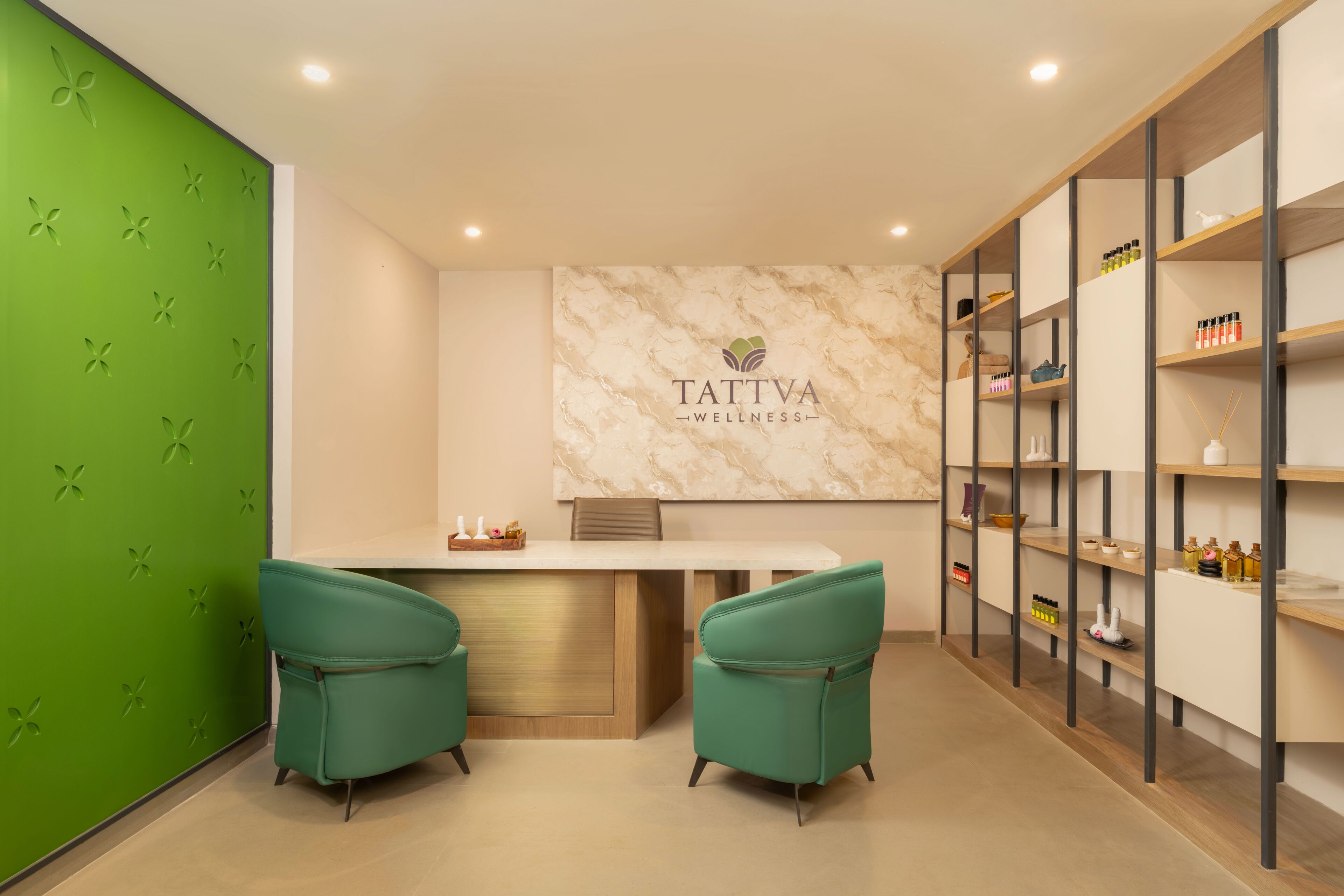 A wellness center with a reception desk, two green chairs, a marble wall with the logo 'TATTVA WELLNESS', and shelves with various items.