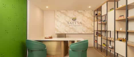 A wellness center with a reception desk, two green chairs, a marble wall with the logo 'TATTVA WELLNESS', and shelves with various items.