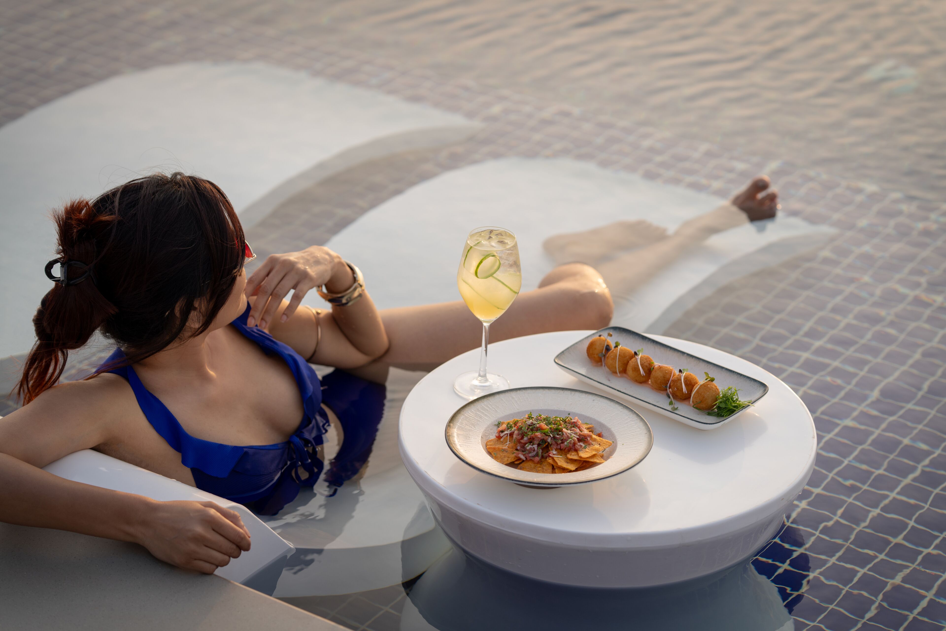 A person relaxing by a poolside table with a plate of food and a drink.