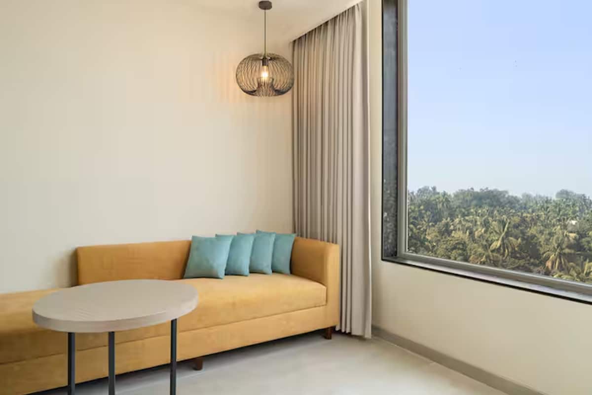 A room with a sofa, a round table, a hanging light fixture, and a large window with a view of palm trees.