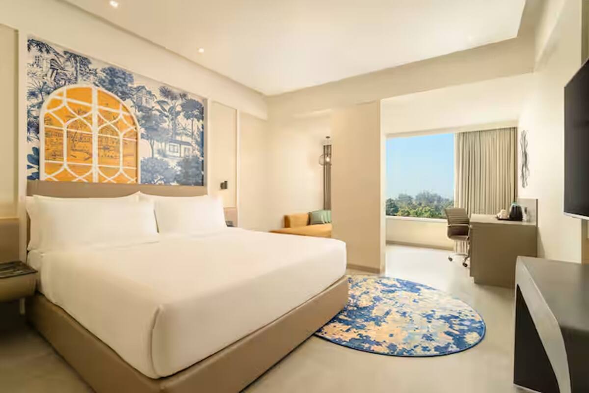 A hotel room with a large bed, a desk, a chair, a TV, and a balcony with a view.