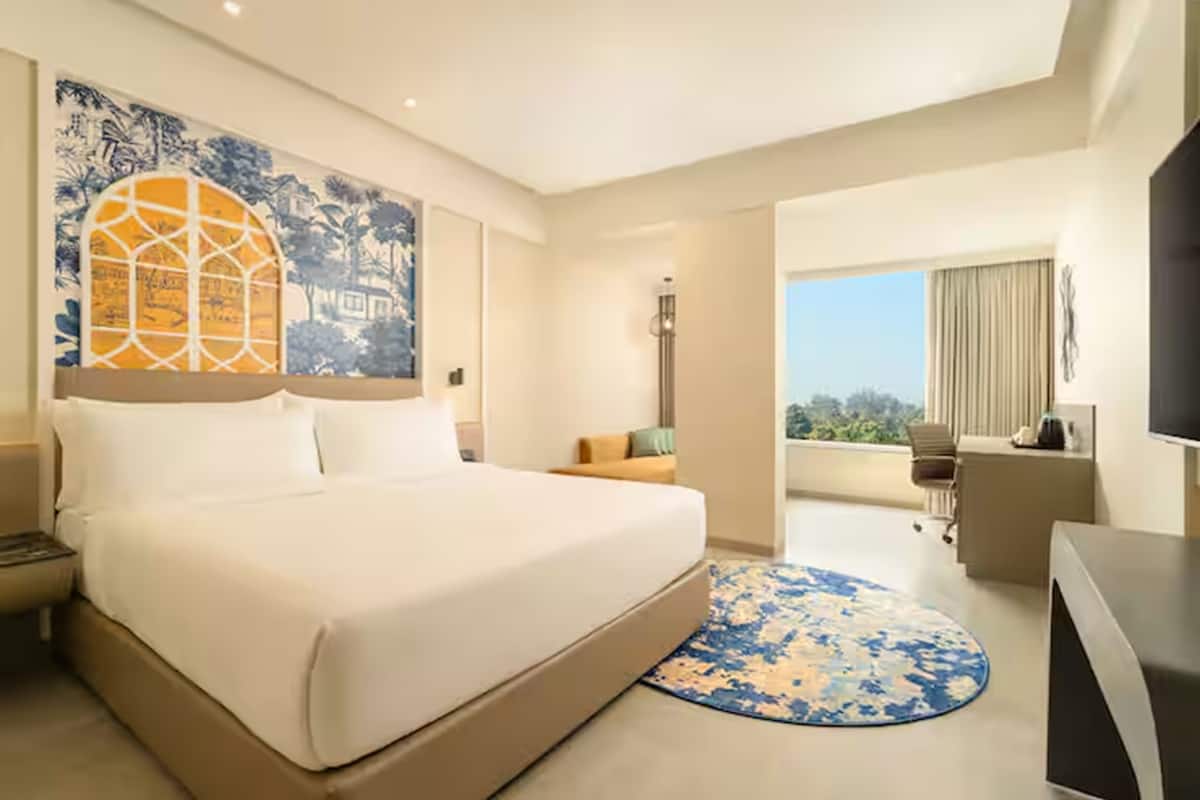 A hotel room with a large bed, a desk, a chair, a TV, and a balcony with a view.