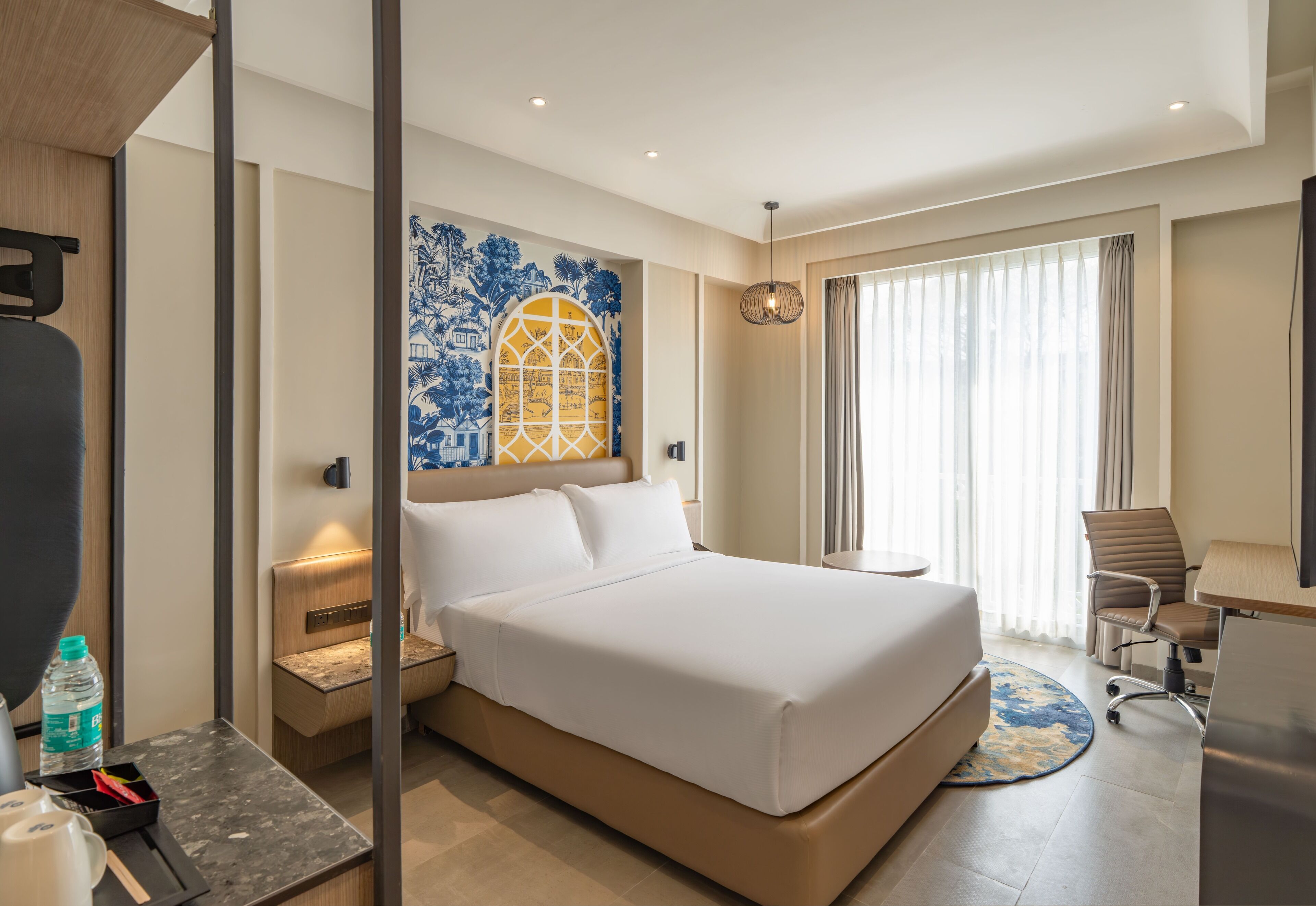 Ein Hotelzimmer mit einem großen Bett, einem Schreibtisch, einem Stuhl, einem kleinen Tisch, einer Wasserflasche und einer Wanddekoration.