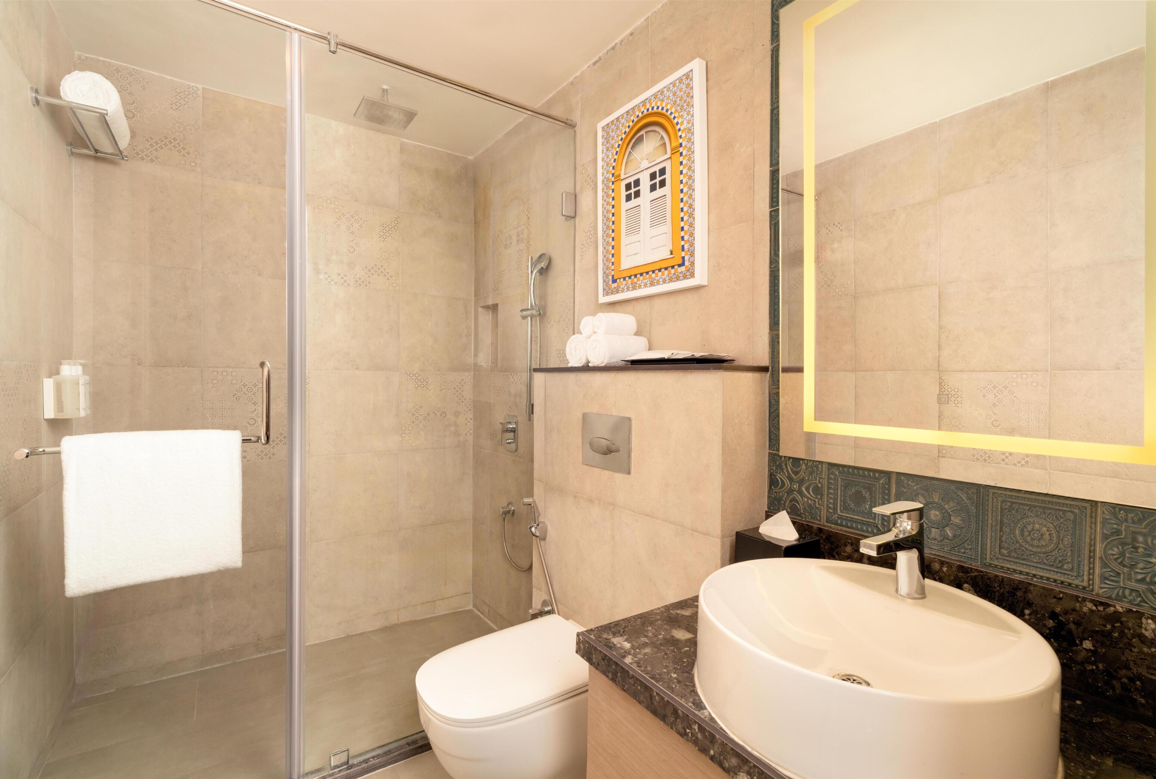 A bathroom with a glass-enclosed shower, a white toilet, a sink with a marble countertop, and a mirror with a decorative tile border.