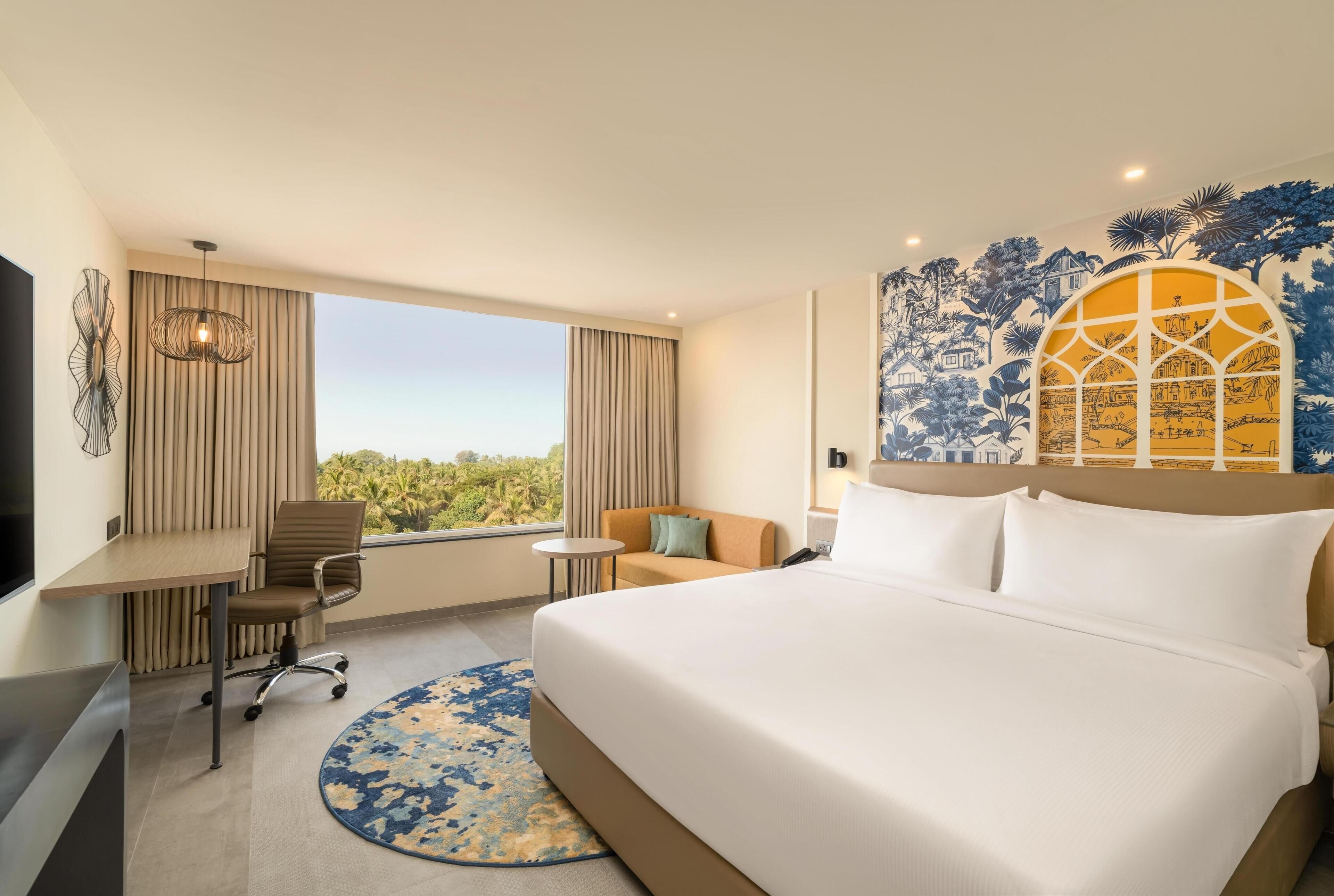 Een hotelkamer met een bed, een bureau, een stoel, een bijzettafeltje, een rond tapijt, een televisie en een groot raam met gordijnen.