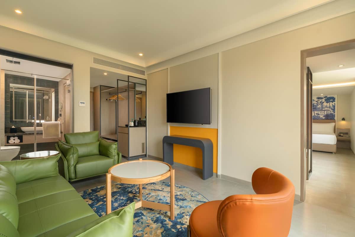 A modern hotel room with a green sofa, an orange chair, a small round table, a flat-screen TV mounted on a wall, and a bathroom with a glass shower enclosure.