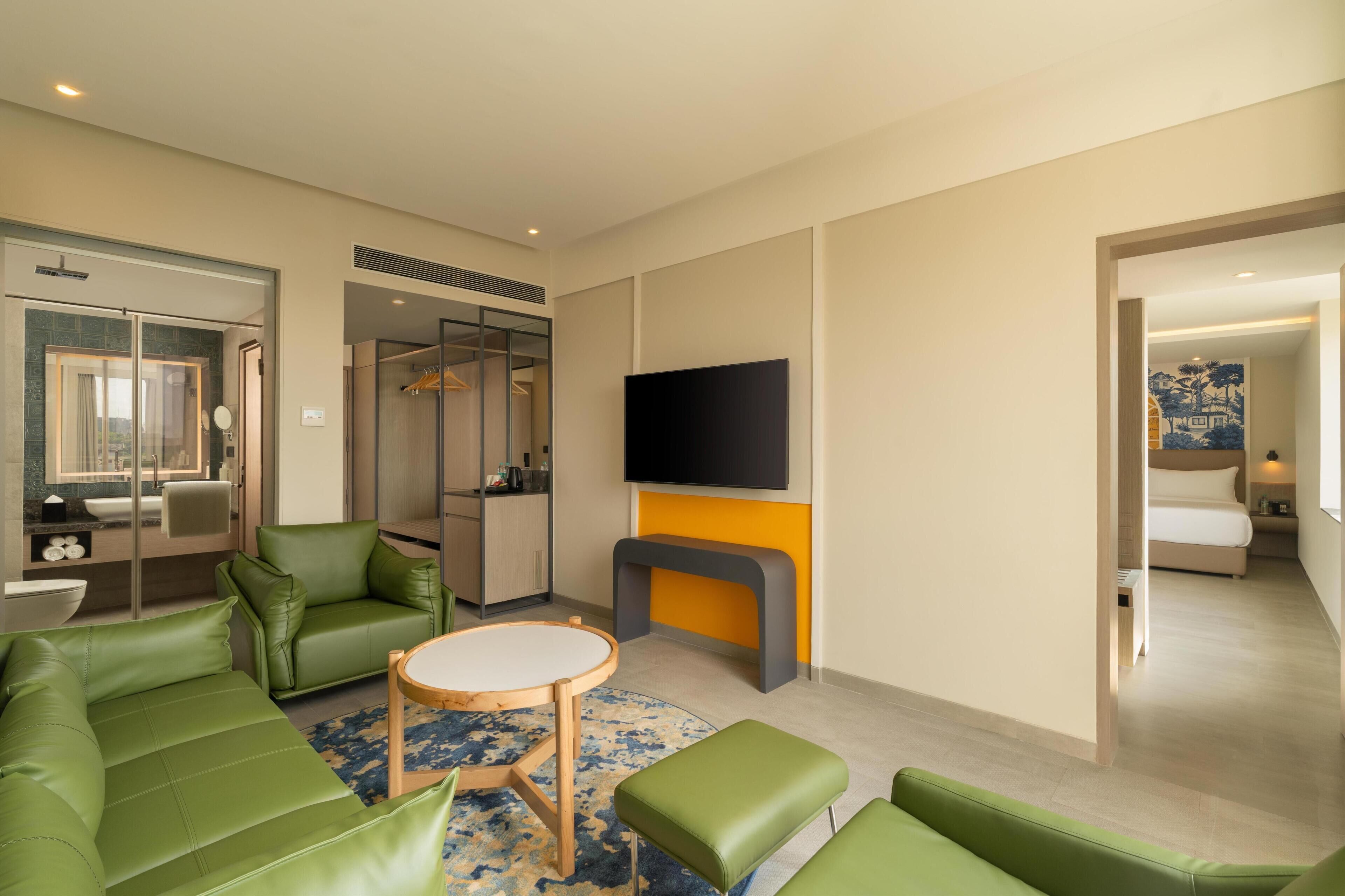 Ein modernes Hotelzimmer mit einem grünen Sofa, einem runden Holztisch, einem an der Wand befestigten Flachbildfernseher und einem separaten Schlafbereich mit Bett und Nachttisch.