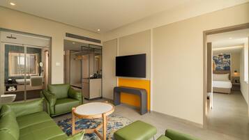 Ein modernes Hotelzimmer mit einem grünen Sofa, einem runden Holztisch, einem an der Wand befestigten Flachbildfernseher und einem separaten Schlafbereich mit Bett und Nachttisch.