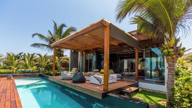 Un área de piscina moderna con terraza de madera, mobiliario y un porche cubierto.