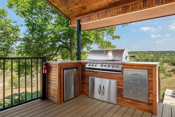 An outdoor kitchen with a grill, a refrigerator, and storage cabinets, set on a wooden deck with a view of trees and hills.