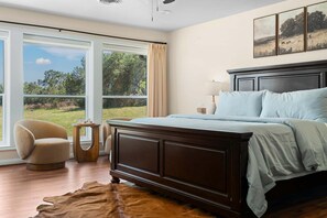 Quarto com uma cama grande, uma cadeira, uma mesinha e vista para uma área gramada através de amplas janelas.