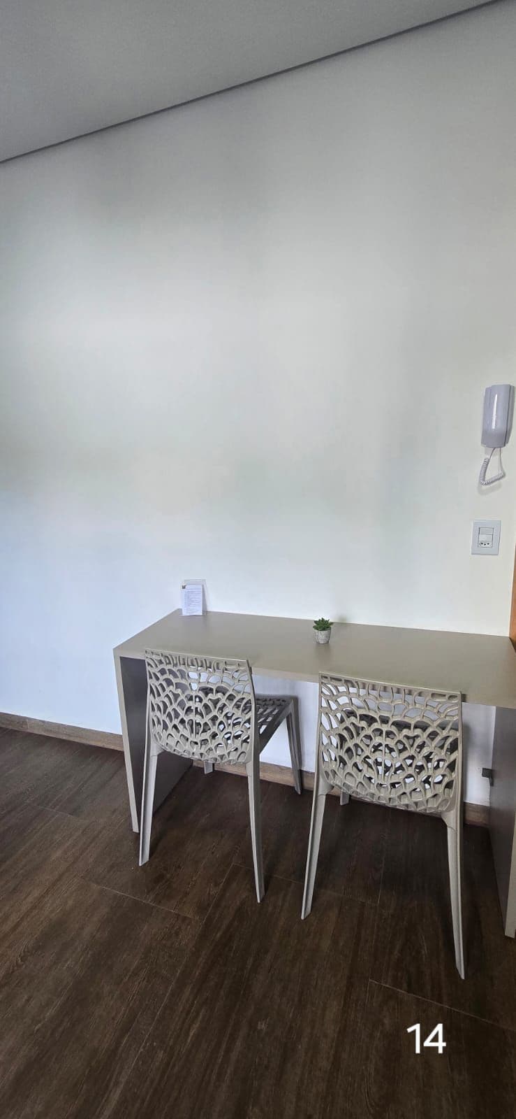 Una habitación pequeña y moderna con una mesa de color claro y dos sillas de respaldo elaborado. Hay una planta pequeña en maceta sobre la mesa, y un teléfono y un interruptor de luz instalados en la pared.