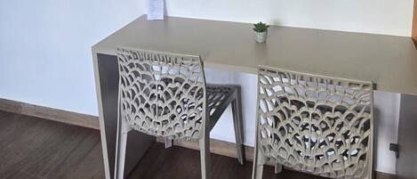 Una habitación pequeña y moderna con una mesa de color claro y dos sillas de respaldo elaborado. Hay una planta pequeña en maceta sobre la mesa, y un teléfono y un interruptor de luz instalados en la pared.