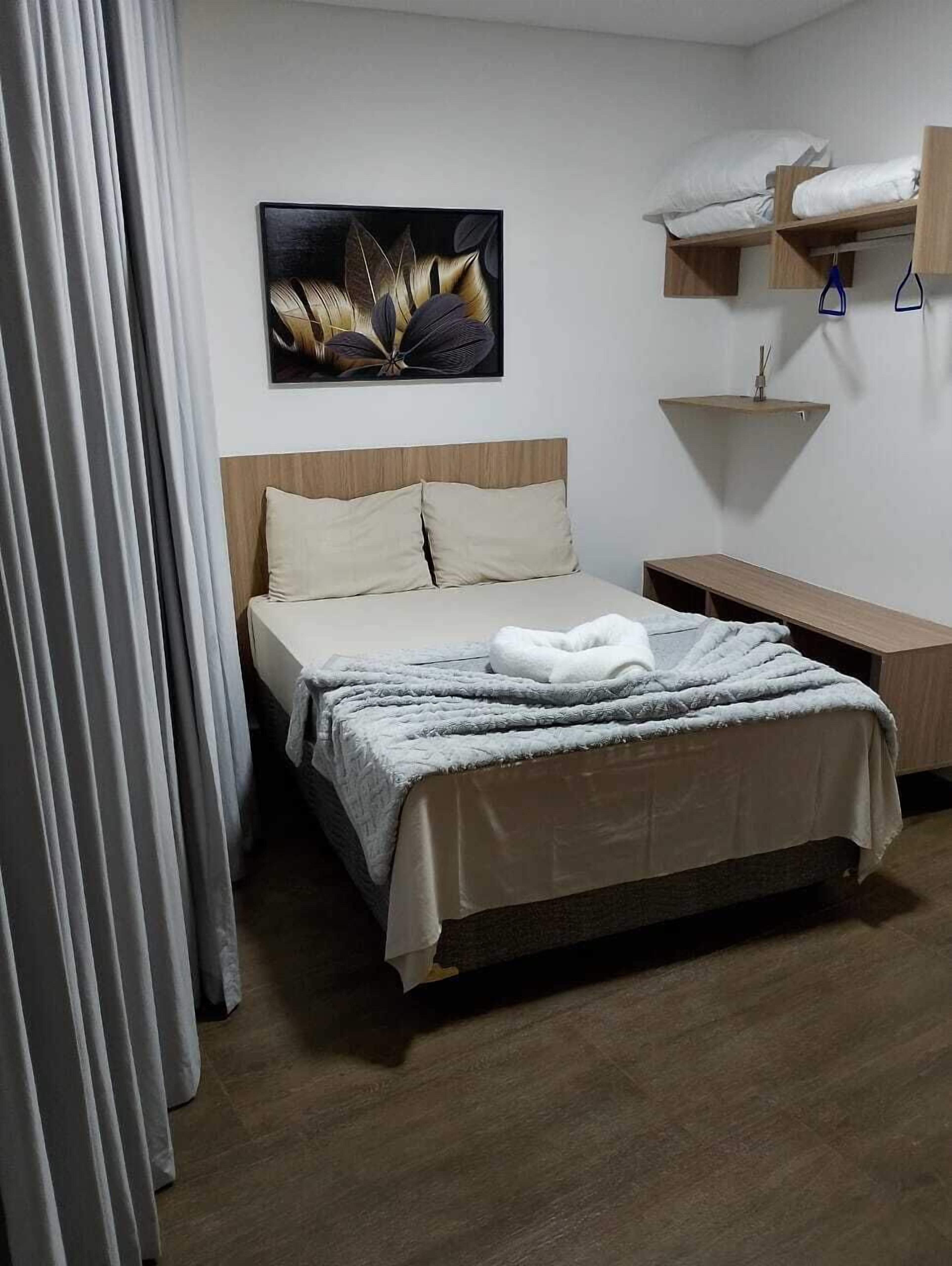 Habitación de hotel ordenada con cama, mesita de noche, un estante pequeño con toallas y una obra de arte colgada en la pared.