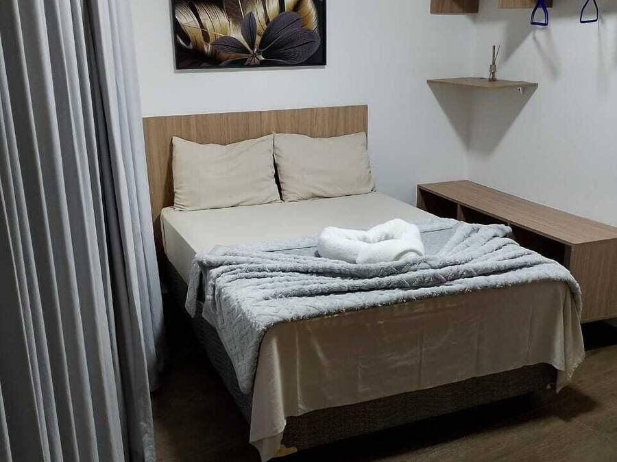 Habitación de hotel ordenada con cama, mesita de noche, un estante pequeño con toallas y una obra de arte colgada en la pared.