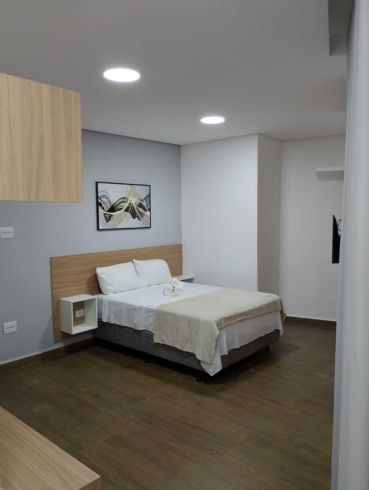 A modern hotel room with a wooden headboard, a neatly made bed, a small bedside table, and a framed abstract painting on the wall.