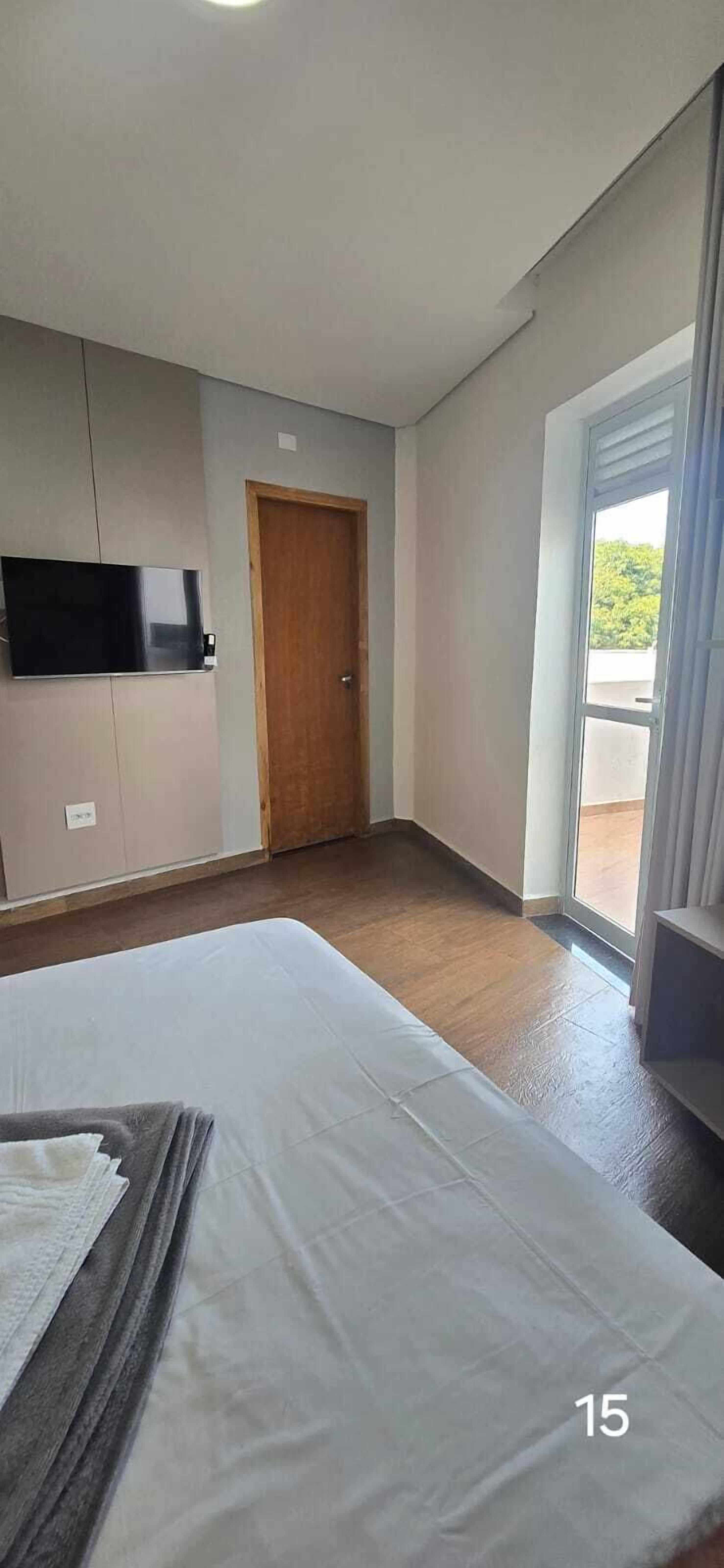 Habitación de hotel con una cama, un televisor montado en la pared, una puerta de madera y un ventanal amplio.