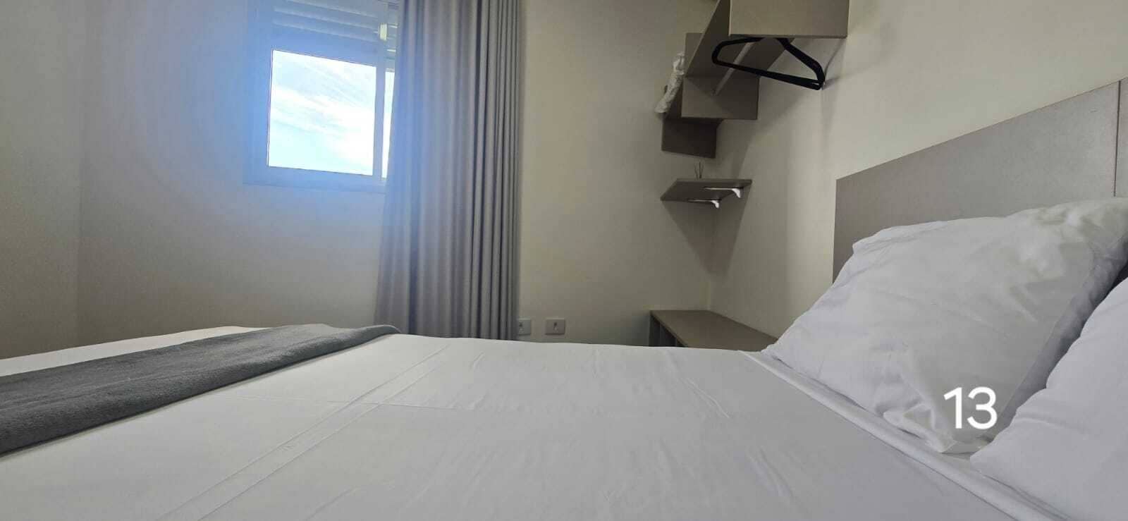 A hotel room with a bed, a nightstand, a hanging coat rack, and a window with curtains.