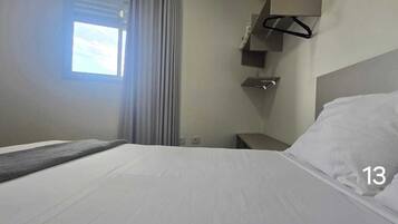 A hotel room with a bed, a nightstand, a hanging coat rack, and a window with curtains.