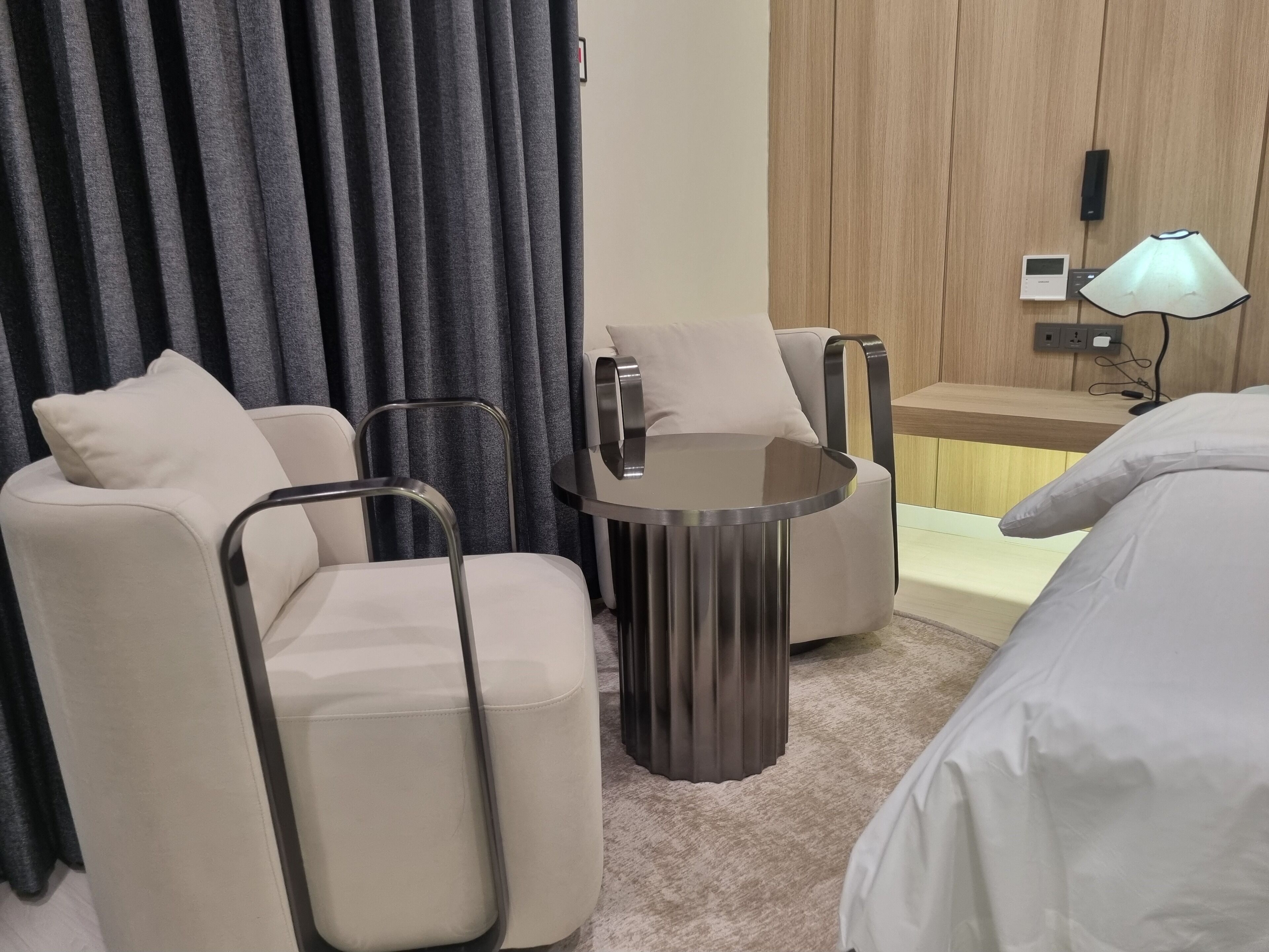 Una habitación de hotel moderna con una cama, una mesa redonda, dos sillones, un sofá, una lámpara y un panel de control en la pared.