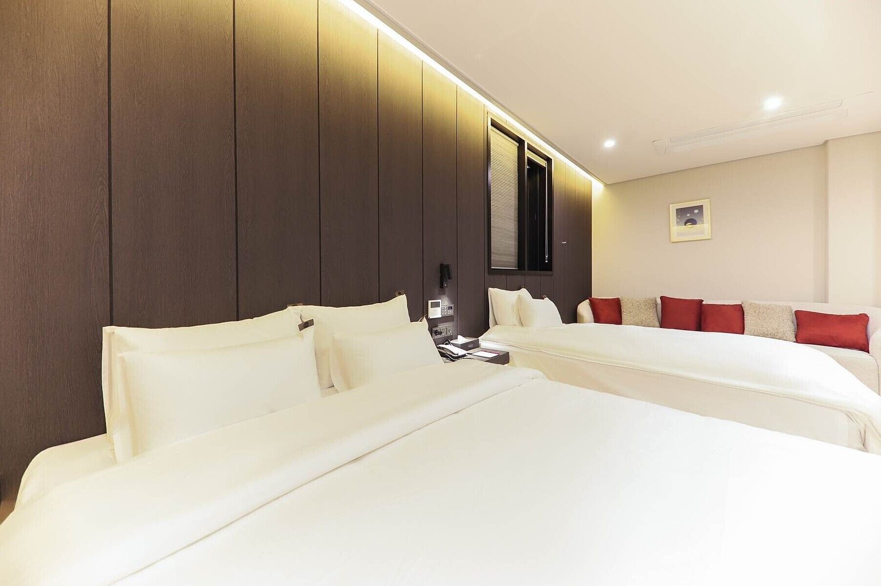 A hotel room with two beds, a wooden panel wall, a bedside table with a phone, and a red and white sofa.
