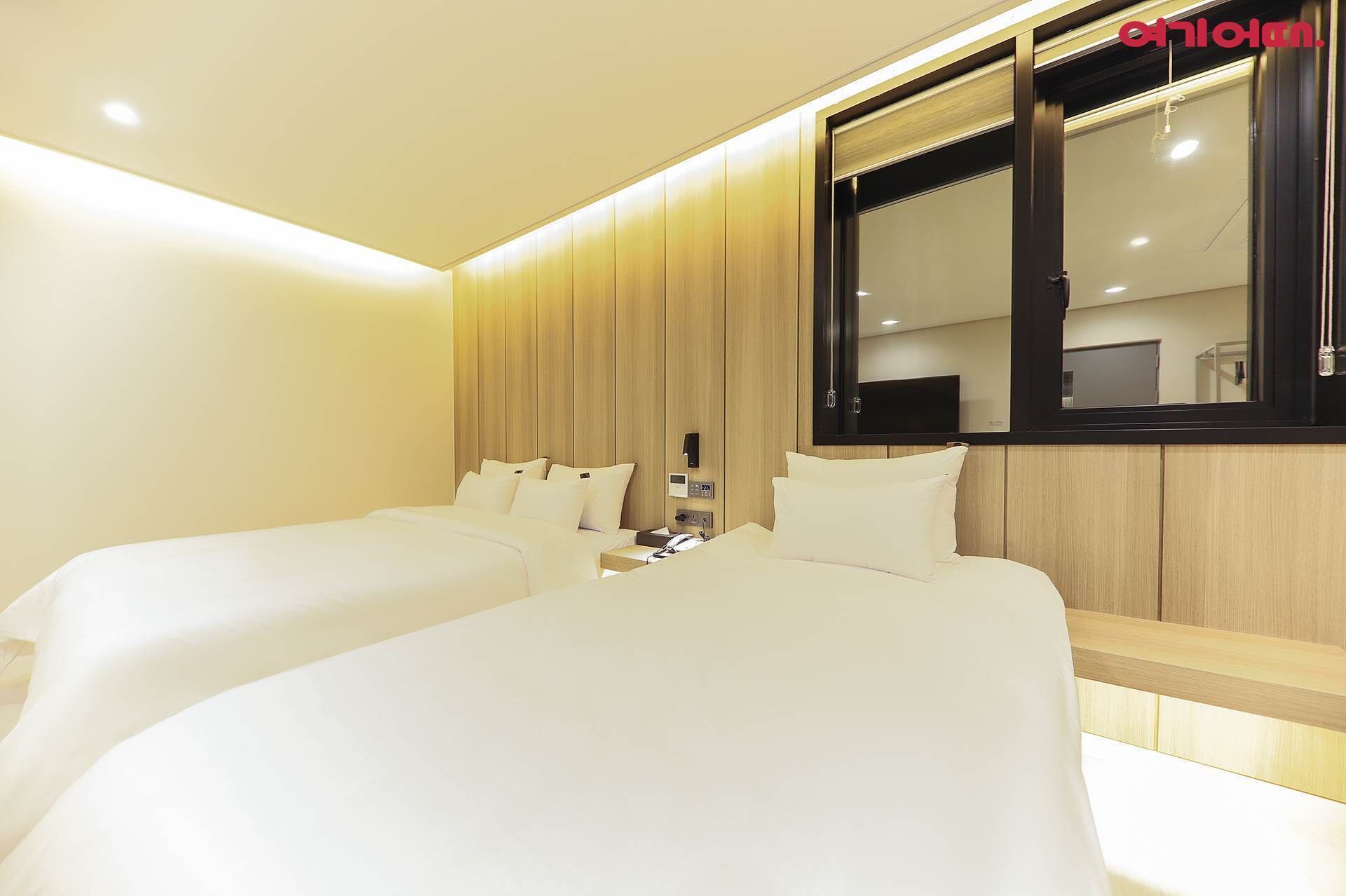 A hotel room with two beds, a wooden panel wall, a large window, and a flat-screen TV.