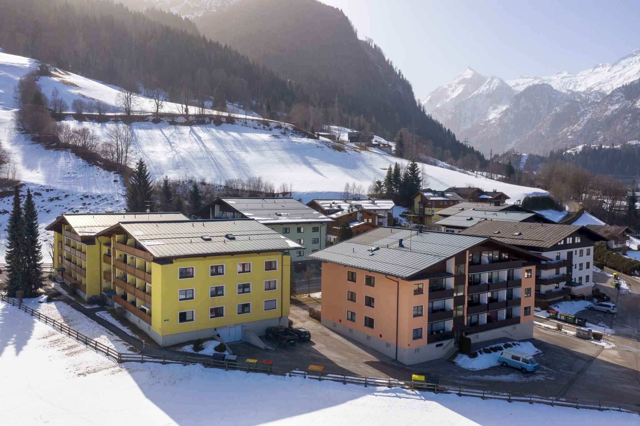 A snow-covered mountain resort with multi-story buildings, a parking area, and a ski slope in the background.