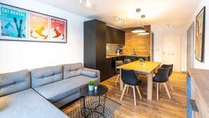 Ein modernes Wohnzimmer mit einem grauen Sofa, einem Holzespeisetisch mit schwarzen Stühlen und einer Küchenzeile mit dunklen Schränken und einer Holzrückwand.