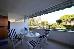 Ein Balkon mit rundem Tisch und gestreiften Stühlen, mit Blick auf einen Garten mit Bäumen und einer Hecke.